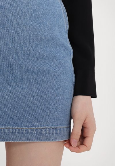 Light blue denim mini skirt with a hemline at mid-thigh, frayed edge, and subtle stitching. Hand resting on the hem, black sleeve visible.