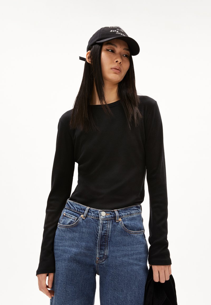 Schwarzes Langarmoberteil, kombiniert mit hochgeschnittenen blauen Jeans. Das Modell trägt außerdem eine schwarze Mütze mit einem weißen Textdesign.