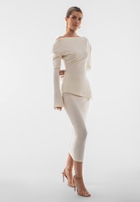 Top a spalle scoperte in maglia a coste color crema, abbinato a una gonna midi a coste dello stesso colore. Presenta maniche lunghe e una silhouette femminile.