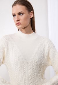 Pull en maille torsadée couleur crème avec un col montant et un design en V. Présente des motifs texturés et des accents côtelés au niveau des poignets et du col.