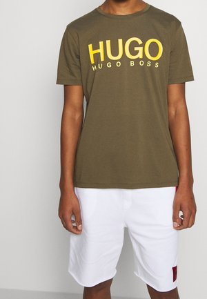 Osoba nosząca oliwkową koszulkę z żółtym napisem "HUGO HUGO BOSS" oraz białe szorty z czerwonym logo na lewej nogawce.