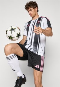 Tenue de football noir et blanc avec des accents roses, présentant des rayures verticales. Le joueur équilibre un ballon de soccer sur son genou, portant des crampons noirs et des chaussettes blanches.