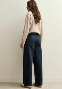 Suéter beige con puños acanalados, combinado con pantalones de mezclilla oscuros de corte holgado que tienen bolsillos traseros y una textura suave.