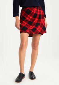 Punainen ja musta ruutuhame, joka on valmistettu pehmeästä kankaasta, ja siinä on kietaisumalli ja hieman levenevä helma, yhdistettynä mustiin kenkiin.