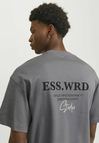 Grå bomulds t-shirt med korte ærmer, en rund halsudskæring og sort grafisk tekst på ryggen, herunder "ESS.WRD." og "Studio."