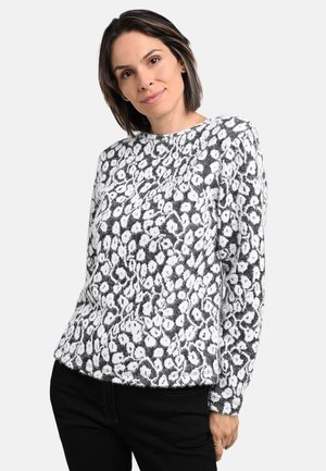 Woman with short dark hair wearing a black and white leopard print sweater and black pants, standing against a plain background.