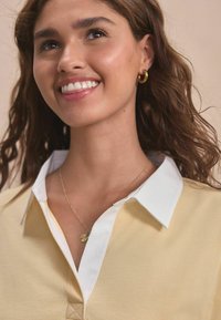 Light yellow polo shirt with a white collar, featuring a small embroidered detail at the neckline and a delicate gold necklace with a round pendant.