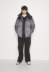 Giacca a vento grigia con una sezione superiore nera, zip frontale e grande colletto. Indossata sopra un hoodie nero e abbinata a pantaloni neri e scarpe da ginnastica.