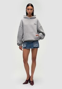 Felpa grigia over-size con tasca frontale, abbinata a una mini gonna di jeans blu con due grandi tasche. La modella indossa scarpe con punta color borgogna.