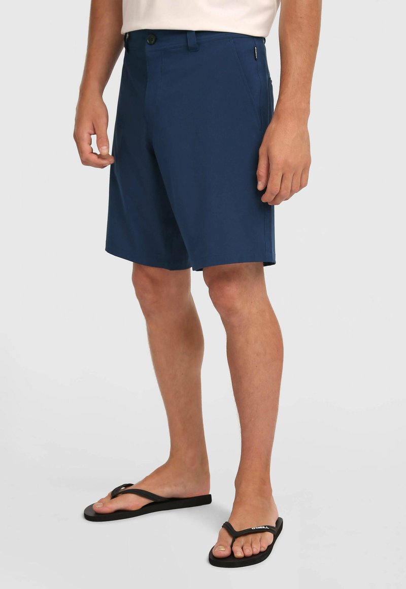 Shorts bleu marine en tissu léger, avec fermeture à bouton, poches latérales et coupe droite, assortis à des tongs noires.