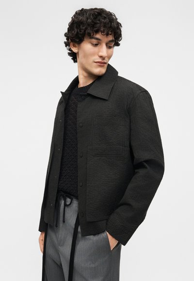 Fiatal férfi hullámos hajjal, fekete texturált gombos dzsekit, fekete kötött pulóvert és szürke húzózsinóros nadrágot visel, lefelé és oldalra néz.