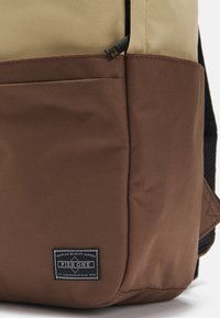 Zaino texturizzato marrone e beige con una toppa nera; presenta un design bicolore e una chiusura con zip per l'accesso.