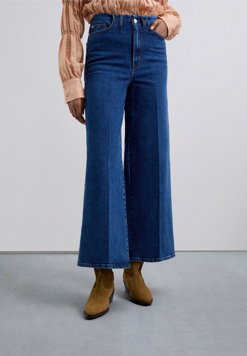 Person som bär högmidjade vida blåjeans, tan mocka ankellånga stövlar och en persikofärgad blus med lång ärm och skrynkliga ärmar.