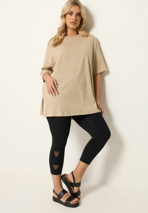 Kvinde iført oversized beige t-shirt, sorte cropped leggings med hjerteudskæringer nær læggene og sorte platform-sandaler, stående op ad hvid væg.