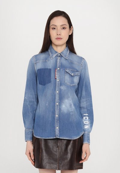 Lichtblauwe denimshirt met een borstzak, voorzien van witte letteraccenten op de mouw en een iets vervaagde textuur. Met knoopsluiting.