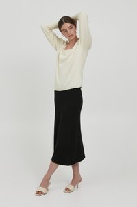 Cremfarbener Langarmtop mit quadratischem Ausschnitt, kombiniert mit einem schwarzen knöchellangen Rock und beigen Absatzschuhen. Glatte Textur überall.