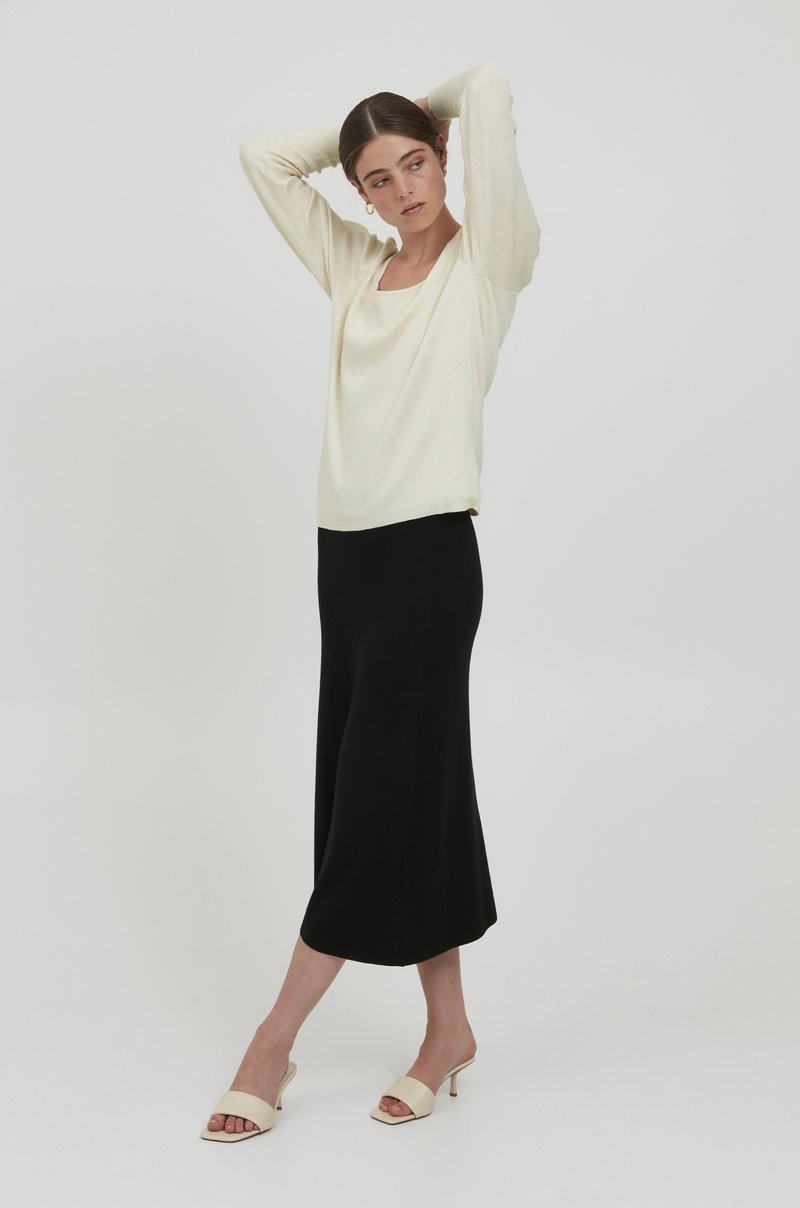 Cremfarbener Langarmtop mit quadratischem Ausschnitt, kombiniert mit einem schwarzen knöchellangen Rock und beigen Absatzschuhen. Glatte Textur überall.