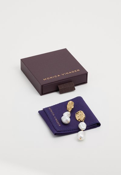 Goldfarbene Ohrringe mit strukturierten Metallakzenten und zwei weißen Perlen, präsentiert in einem lila Stoffbeutel innerhalb einer dunkelpurpurnen Box.
