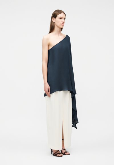 Nő, aki tengerészkék, aszimmetrikus, egyrészes vállú felsőt visel, hosszú, fehér, magas hasítású maxiszoknyával, fekete pántos magassarkúban áll.