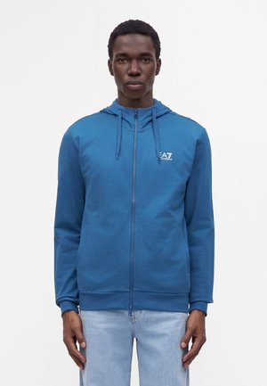 Homme portant un sweat à capuche bleu zippé avec le logo EA7 et un jean bleu clair, debout sur un fond blanc.