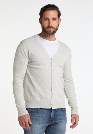 Man met baard die een lichtgrijze gebuttonde cardigan draagt over een wit overhemd en blauwe spijkerbroek, staand tegen een witte achtergrond.