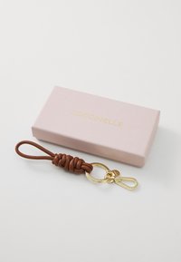 Portachiavi in pelle marrone con design annodato, attaccato a una clip in metallo dorato, accanto a una scatola rosa chiaro con testo dorato.