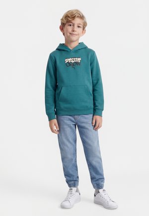 Niño rubio de pie con sudadera con capucha teal "Street Crew", jeans azul claro con puños elásticos y zapatillas blancas sobre un fondo liso.