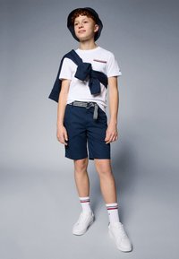 Tengerészkék rövidnadrág, fehér pólóval, amely mellzsebbel és piros/kék csíkokkal díszített, kiegészítve tengerészkék pulóverrel, valamint egy vödörkalappal. Az összeállítást fehér sportcipő teszi teljessé.