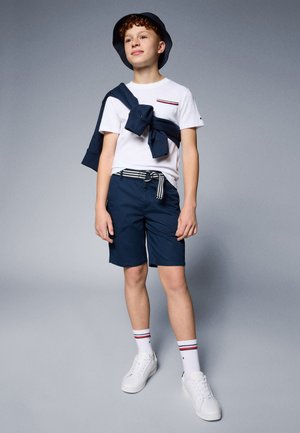 Balta marškinėliai su raudonu ir tamsiai mėlynu juostų akcentu, tamsiai mėlyni šortai, mėlynas megztinis užmestas ant pečių, tamsiai mėlynas kibiro skrybėlė, balti sportbačiai, juostuoti pėdkelniai.