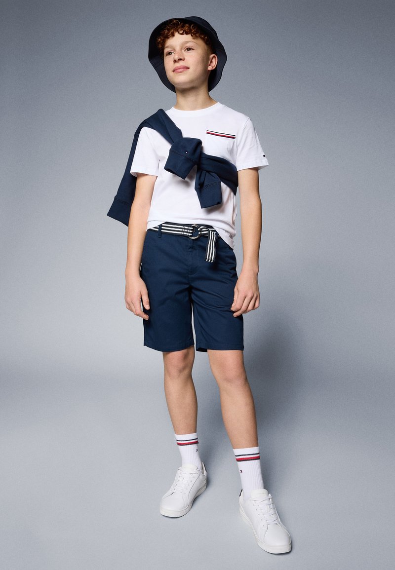 Tengerészkék rövidnadrág, fehér pólóval, amely mellzsebbel és piros/kék csíkokkal díszített, kiegészítve tengerészkék pulóverrel, valamint egy vödörkalappal. Az összeállítást fehér sportcipő teszi teljessé.