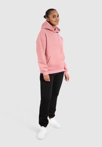 Haut à capuche rose avec une poche avant, des manches retroussées et un cordon de serrage, associé à un pantalon de survêtement noir et des baskets blanches.