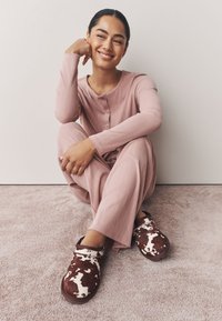 Abbigliamento da casa rosa chiaro a coste con scollatura rotonda. Zoccoli marroni e bianchi con motivo a fantasia. Sfondo in morbido tappeto beige. Ambientazione minimalista.