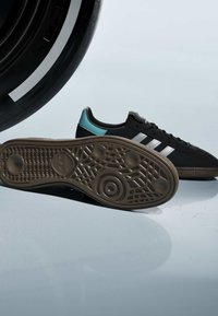 Baskets noires Adidas avec des rayures blanches et un détail turquoise au talon, montrant une semelle marron texturée, sur un fond clair avec un grand pneu noir.
