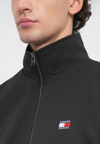 Sort lynlåsjakke med høj krave. Har et lille logo patch i blå, rød og hvid. Glat stof med en mat tekstur.