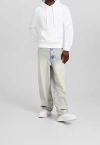 Weiße Kapuzensweatshirt mit einer Fronttasche, kombiniert mit hellblauen, weiten Jeans. Weiße Sneaker runden das lässige Outfit ab.