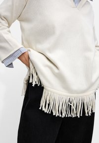 Sweter w kolorze kremowym, robiony na drutach, z dekoltem w serek, z długimi frędzlami u dołu, noszony na niebieskiej i białej koszuli w paski.