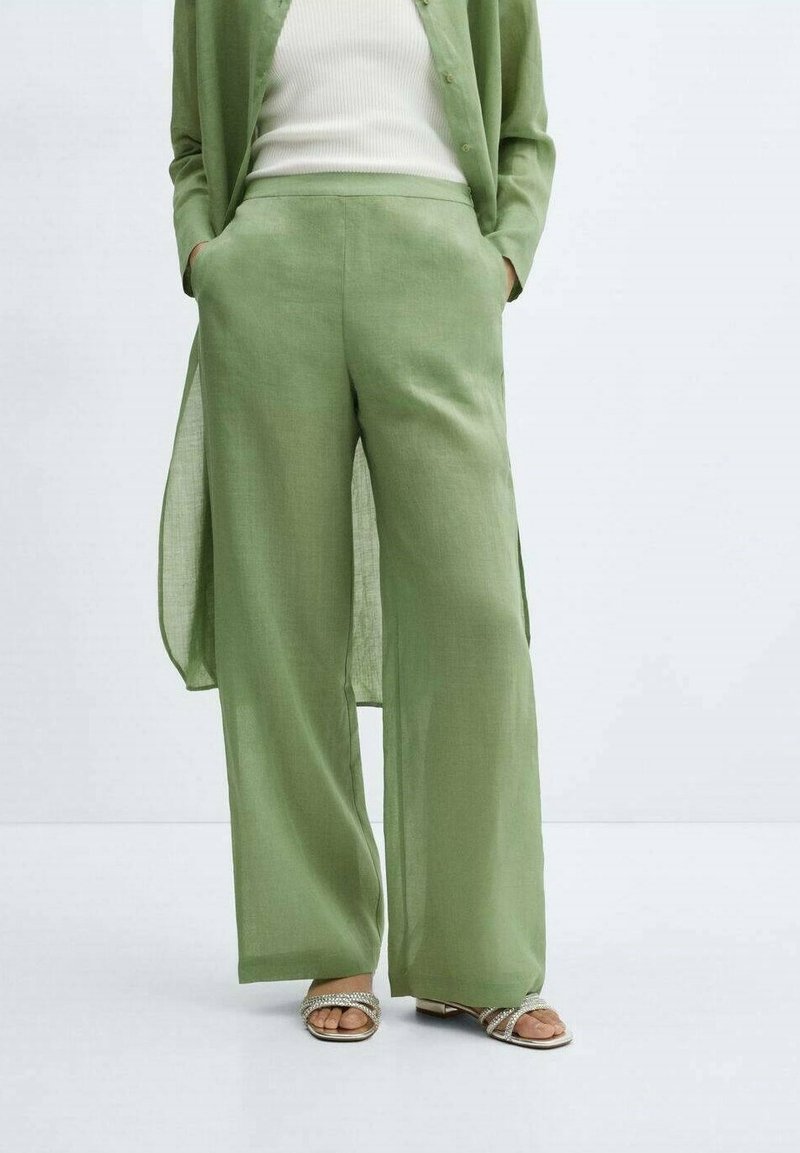 Femme portant un pantalon large vert clair et un long cardigan assorti sur un haut blanc à côtes, posant les mains dans les poches.