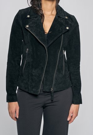 Woman wearing black suede biker jacket with asymmetric zipper and silver buttons, paired with black pants, standing against gray background.