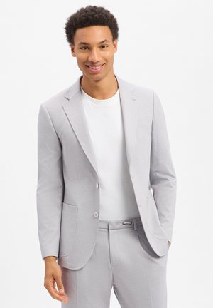 Junger Mann lächelt, trägt hellgraues Sakko und passende Hose über einem weißen T-Shirt, steht vor weißem Hintergrund.