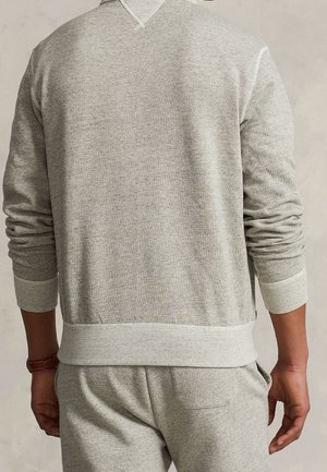 Achteraanzicht van een persoon die een lichtgrijze sweater met lange mouwen en bijpassende joggingbroek draagt, met de handen ontspannen langs het lichaam.
