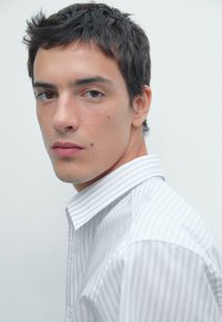 Camicia bianca con righe grigie sottili, dotata di colletto a punta e vestibilità rilassata. Tessuto morbido, prospettiva semi-girata.