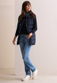Marineblaue Pufferweste mit hohem Kragen, Reißverschluss und Kordelzug. Getragen über einem schwarzen Oberteil und kombiniert mit hellblauen Jeans und weißen Sneakers.
