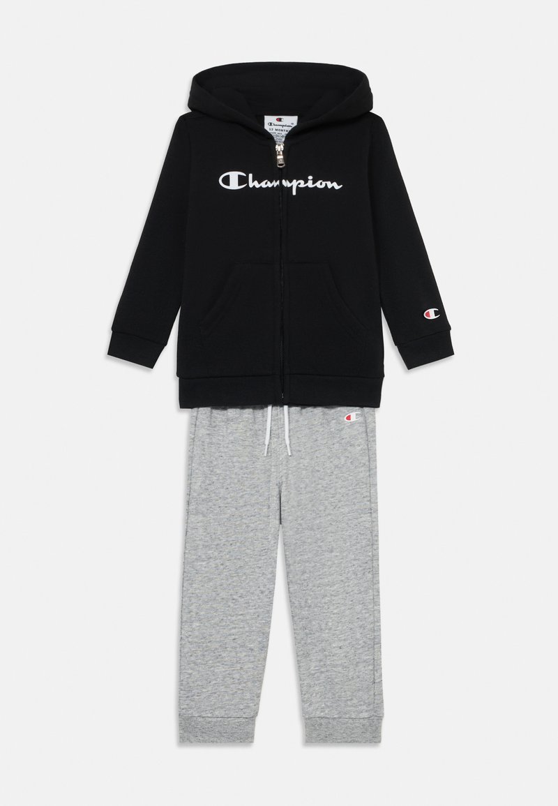 Melnas kapuces blūze ar baltu "Champion" logotipu un priekšējām kabatām, kombinēta ar pelēkiem treniņbikšu, kam ir elastīga josta un aproces.