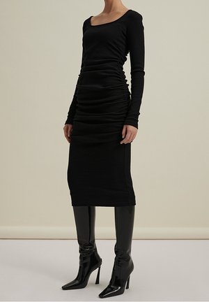 Robe noire à manches longues avec un décolleté carré et des détails froncés. Texture lisse avec une silhouette ajustée, accompagnée de bottes noires brillantes jusqu'aux genoux.
