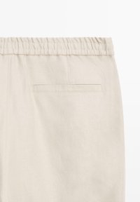 Lichtbeige broek met een elastische tailleband en een enkele horizontale achterzak zonder zichtbare sluiting.
