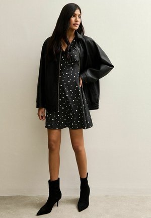 Zwarte leren bomberjack over een zwarte jurk met een witte sterrenprint, gecombineerd met zwarte enkellaarzen. De gladde texturen en de getailleerde pasvorm zijn duidelijk zichtbaar.