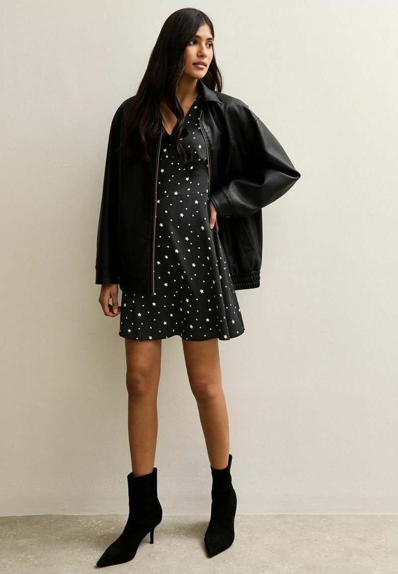 Zwarte leren bomberjack over een zwarte jurk met een witte sterrenprint, gecombineerd met zwarte enkellaarzen. De gladde texturen en de getailleerde pasvorm zijn duidelijk zichtbaar.
