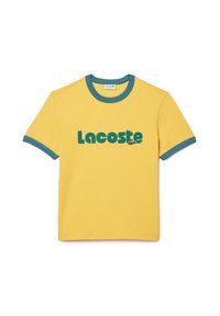 Żółta bawełniana koszulka z niebieskimi ściągaczami przy dekolcie i rękawach, z wyraźnym zielonym logo "Lacoste" nadrukowanym z przodu.