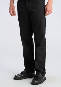 Jeans in denim nero con vestibilità dritta, caratterizzati da una texture liscia e design a cinque tasche. Abbinati a mocassini neri con dettagli a nappina.