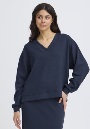 Femeie cu păr lung și închis la culoare, purtând un pulover larg cu decolteu în V, de culoare bleumarin, și o fustă asortată, pozând cu o mână pe șold.